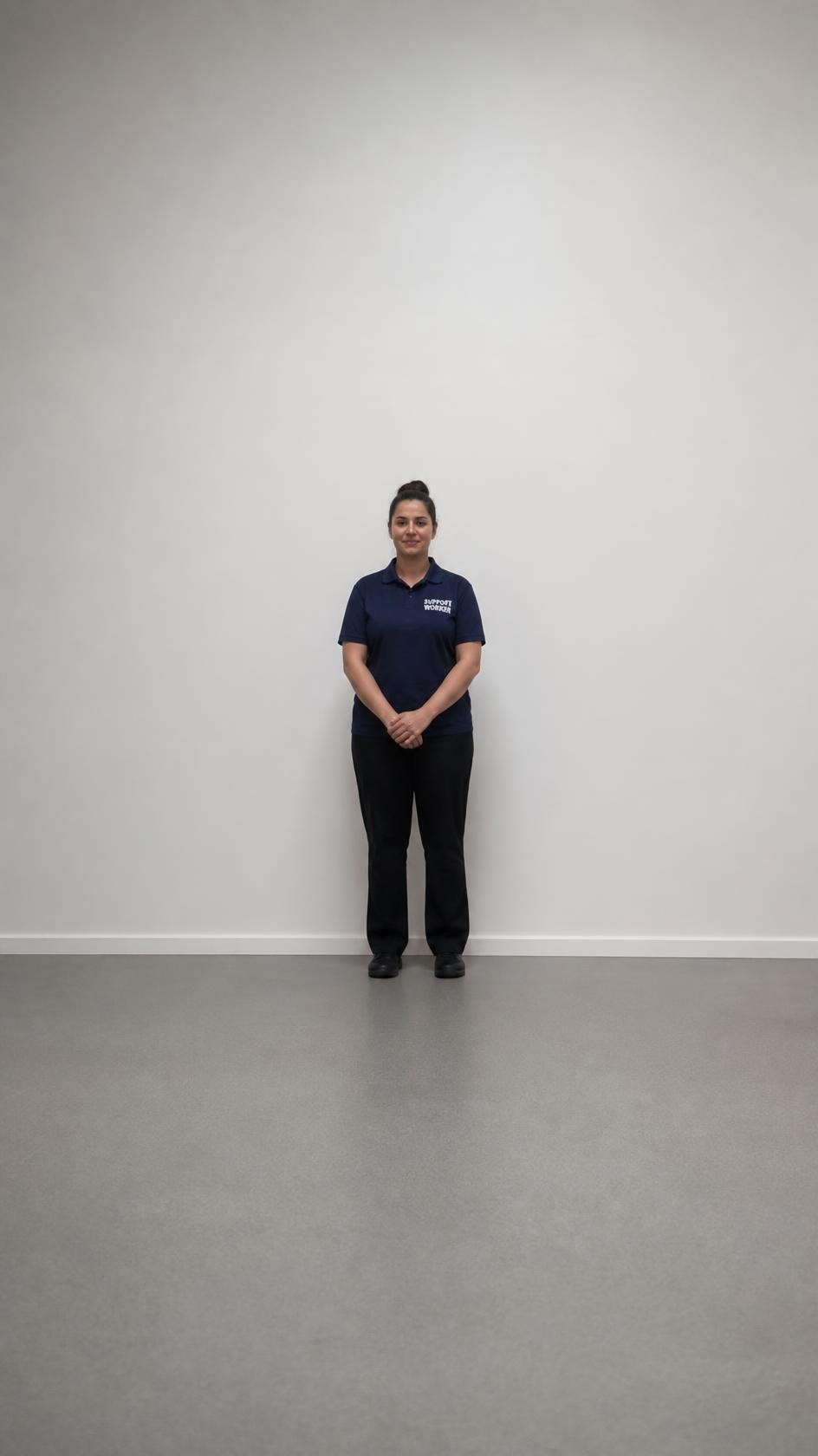 A full-body shot of a support worker standing against a plain wall.